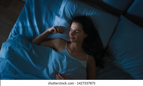 Blick von oben auf die schöne junge Frau, die nachts gemütlich auf einem Bett in seinem Schlafzimmer schläft. Blaue nächtliche Farben mit kaltem, schwachem Laternenlicht, das durch das Fenster scheint.