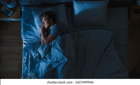 Draufsicht auf einen hübschen jungen Mann, der nachts gemütlich auf einem Bett in seinem Schlafzimmer schläft. Blaue nächtliche Farben mit kaltem, schwachem Laternenlicht, das durch das Fenster scheint.