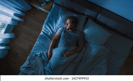Draufsicht des hübschen jungen Mannes, der gemütlich auf einem Bett in seinem Schlafzimmer nachts schläft. Blaue nächtliche Farben mit kaltem, schwachem Laternenpfahllicht, das durch das Fenster scheint.