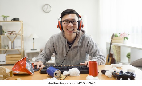 vista frontal de un joven universitario juguetón jugando videojuegos en un apartamento desordenado en las vacaciones de verano en casa. chico hogareño sonriendo mirando a la cámara mientras la pantalla de la computadora usa auriculares emoción emocionada.