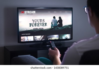 Online movie stream service in smart tv. Streaming series with on demand video (VOD) service in television. Man choosing film to watch with remote. Person sitting on couch at home late at night.