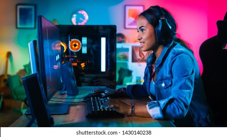Portrait of a Pretty and Excited Black Gamer Girl with Headphones Smiling to the Camera. She is Sitting on Chair next to Powerful Personal Computer. Home and PC is Lit with Neon Lights.