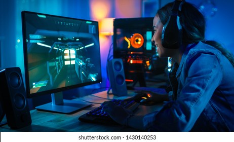 Hübsches und aufgeregtes schwarzes Blogger-Mädchen in Kopfhörern spielt Ego-Shooter-Online-Videospiel auf ihrem Computer. Raum und PC haben bunte Neon-LED-Lichter. Gemütlicher Abend zu Hause.