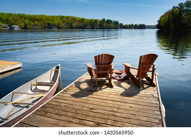 Two Adirondack chairs on a wooden dock facing the blue water of a lake in Muskoka, Ontario Canada. Life jackets are visible near the chairs. A canoe is tied to the pier, paddles are stored inside.
