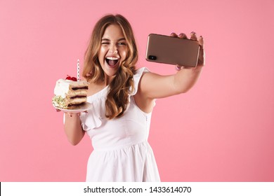 携帯電話で自撮り写真を撮り、ピンクの背景に分離されたろうそくでケーキを保持しながらウィンクしている若い笑いの女性の画像