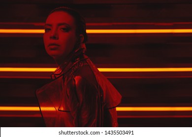 Chân dung phong cách Cyberpunk của cô gái trong chiếc áo khoác bóng kính và chiếc váy đỏ của tương lai. Cô ấy tạo dáng dựa vào bức tường đèn neon. Thiết lập được thắp sáng bằng ánh sáng đỏ. Hình ảnh có tông màu tối. Tường neon có màu đỏ