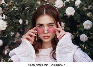 Nahaufnahme im Freien, Porträt einer jungen, schönen Dame mit rosafarbener, runder Sonnenbrille, weißer Vintage-Bluse, Model mit Blick in die Kamera, posiert im blühenden Rosengarten