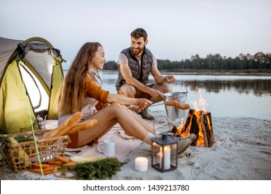Pasangan muda dan ceria memasak sosis di perapian, berpiknik di perkemahan di pantai pada malam hari