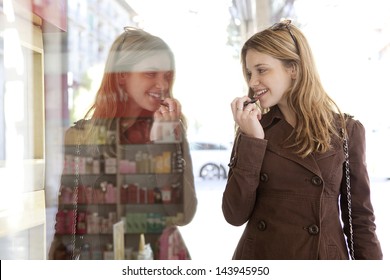 Close up side portrait of a young teenager tourist visiting the city and applying lipstick using the reflection of a shopping mall cosmetics store window.