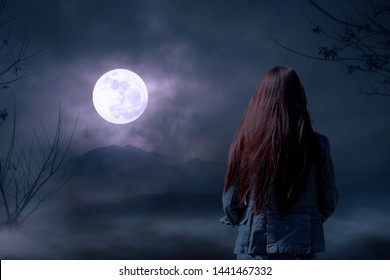 雲のある夜空に対する乾いた木々のシルエットと、夜だけで満月を見る女性スタンド、自然の背景、暗いトーン