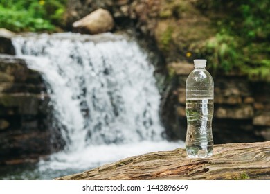 Garrafa de plástico transparente com água fica em um tronco de madeira contra o fundo do rio e da cachoeira. O conceito de água potável natural pura
