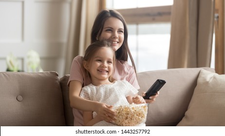 Happy mother with cute little daughter watching tv, eating popcorn snack, using remote controller, sitting on couch together, having fun, cute preschool girl sitting on smiling mum knees at home