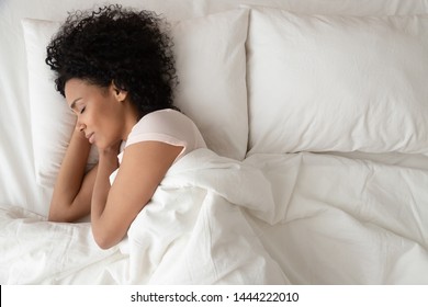 Gelassene, ruhige Afroamerikanerin, die in einem bequemen Bett schläft, das auf einer orthopädischen Matratze mit weichem Kissen liegt, friedliche junge schwarze Dame, die mit einer Decke bedeckt auf weißen Laken im Schlafzimmer ruht, Draufsicht
