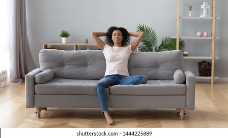 Young calm black woman relaxing sit on comfortable sofa in modern living room, lazy happy african woman girl resting on couch breathing fresh air enjoy peace of mind no stress free on couch at home