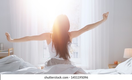 Frumoasa brunetă se trezește dimineața, se întinde în pat, soarele strălucește pe ea de la fereastra mare. O tânără fericită salută ziua nouă cu lumina caldă a soarelui.