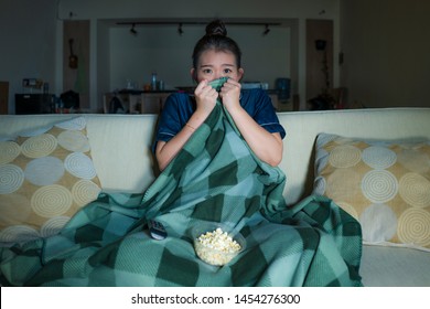امرأة كورية آسيوية شابة جميلة خائفة ومرعوبة تشاهد فيلم رعب أو فيلم إثارة تأكل الفشار في خوف وتبدو على وجهها وهي تأكل الفشار وتجلس على أريكة غرفة المعيشة في الظلام