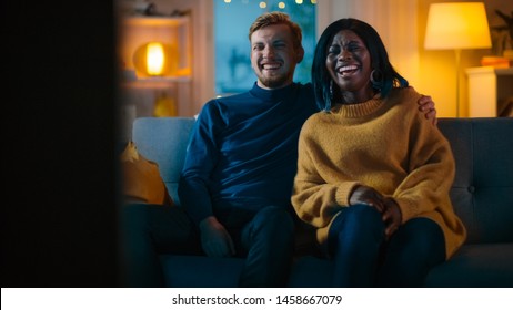 Gelukkig divers jong stel kijken naar komedie op tv terwijl ze op een bank zitten, ze lachen en genieten van de show. Knappe blanke jongen en zwart meisje verliefd tijd samen doorbrengen in het gezellige appartement.