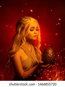 Eine junge blonde Frau hält ein großes, ungewöhnliches Drachenei in den Händen. Sie sieht ihn hoffnungsvoll an. Hintergrund rot flammendes Feuer mit Rauch und Funken. Studio kreative Fotografie Daenerys Targaryen