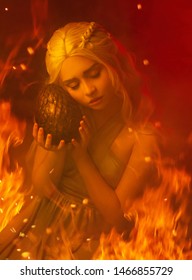 A young blond woman in a gray dress is sitting on fire and hugging a dragon egg. Elven Princess. Red fiery background. Art closeup portrait. Daenerys Targaryen. The scene is the birth of a dragon.