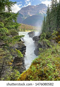 Bergwasserfälle zwischen schroffen felsigen Klippen voller Kiefern, aufsteigendem Wassernebel und Bergen im Hintergrund.