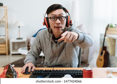 Young man playing game at messy home and streaming walkthrough video. Shouting crazy guy with angry emotion point finger at computer screen. mad boy with bad teammate player online and scolding