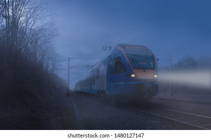 A regional train at night in the moonlight and fog on a free route near the German city of Kassel. You can see masts, rails and power lines and the front lights in the fog.