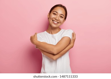 Love yourself. Pretty glad Asian girl embraces herself, feels comfort and care, tilts head, wears white t shirt, has no make up, isolated over rosy wall, thinks about lover, wants be in his warm arms
