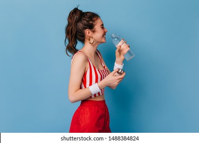 赤い縞模様の衣装を着た若い女の子が笑い、孤立した背景にボトルから水を飲む