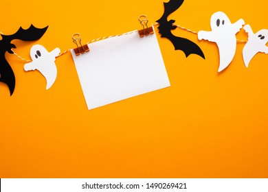 Feliz concepto de halloween. Decoraciones de Halloween, tarjeta de papel en blanco en clips, murciélagos, fantasmas sobre fondo naranja. Maqueta de tarjeta de felicitación de fiesta de Halloween con espacio de copia. Endecha plana, vista superior, arriba.