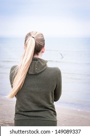 Young woman with long blond ponytail on a foggy morning looking at the sea. There is a gull above the water, but you can see only light shade of it. 