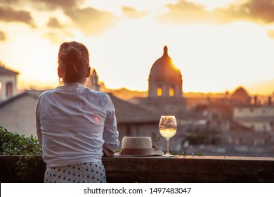 Young woman tourist fashion white dress with glass of white wine in front of panoramic view of Rome cityscape from campidoglio terrace at sunset. Landmarks, domes of Rome, Italy.