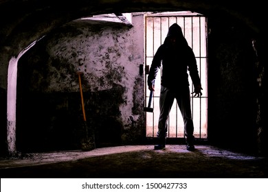 Gruseliger dunkler Mann mit Hammer im Verlies - Silhouette eines Serienmörders, der mit bedrohlicher Haltung in gruseligem Gefängnis steht - Konzept von Wahnsinn und Mord - Gegenlichtbild mit verstärktem Kontrast