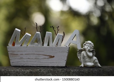 E klenge wäissen Engel nieft enger wäisser Holzkëscht mam Wuert Mama drop.