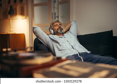 Mann mittleren Alters, der Musik mit Kopfhörern auf dem Phonographen hört, entspannt auf dem Sofa in seinem Haus