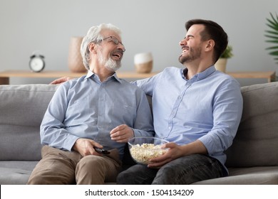 Happy two generations male family laugh enjoy talk watching funny comic tv show, cheerful old senior father and young adult son hold snack remote control bonding relax view television sit on sofa