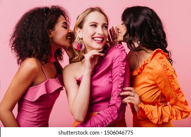 Retrato de estudio de tres amigos internacionales sonriendo sobre fondo rosa. Foto de medio cuerpo de chicas compartiendo noticias.