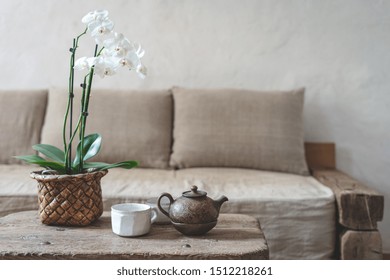 Selektiver Fokus der Ton-Teekanne, der weißen Tasse und der Orchideenblume am Holztisch gegen unscharfe Wand mit Kopienraum auf Hintergrund. Gemütliches Haus mit Komfortsofa im Vintage-Interieurstil