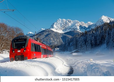 A DB train travels thru a forest in a valley covered by heavy snow & Karwendel mountain stands under clear sky in background on a sunny winter day near Klais & Garmisch-Partenkirchen, Bavaria, Germany