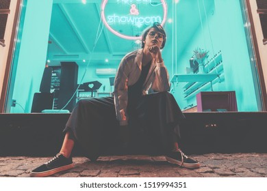 Tomboy teen hipster fashion hairstyle e shkurtër gen z vajzë veshin rroba me stil syzet ulur në rrugë pranë shenjës neoni, modelja femër grua shikon kamerën në qytetin dritë natën dritë stil të viteve 80, portret