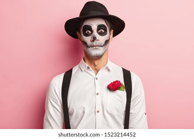 Portret van griezelige knappe man maakte make-up voor Halloween-evenement, heeft afbeelding van vampier of geest, roodroze bloem in zak van wit overhemd, draagt ​​zwarte hoed, heeft een enge blik, gekleed in zombiekleding