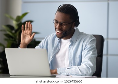 Foto da cabeça de um empresário afro-americano sorridente usando fones de ouvido, fazendo videochamada com clientes, cumprimentando parceiros no escritório. Palestrante inteligente de raça mista dando boas-vindas aos participantes do webinar online.