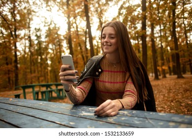  Beautiful girl plays a game on the phone in a autumn park. Different emotions from a phone application. Girl writes a message on a mobile phone.  Woman write message. Lifestyle. Colorful autumn.