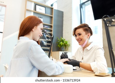 cheerful open-minded manicurist in white clothes make manicure to young caucasian woman with red hair in shirt, Background beauty salon