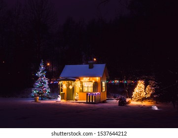 Rumah bermain anak-anak pribadi kuning dicat kayu yang lucu di taman rumah, dihiasi dengan lampu tali LED Natal di luar ruangan di malam penggulung bersalju yang dingin. Pohon cemara Natal yang dihias.