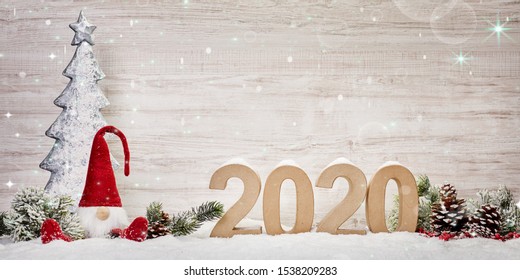the numbers for the year 2020 stand in the snow with a pine branch decoration and a little imp sit on the left site with a christmas tree in the back on a wooden texture background with shining stars