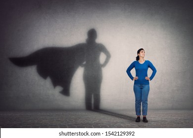 Brave woman keeps arms on hips, smiling confident, casting a superhero with cape shadow on the wall. Ambition and business success concept. Leadership hero power, motivation and inner strength symbol.