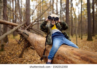 Little boy scout with binoculars during hiking in autumn forest. Child is sitting on large fallen tree and looking through a binoculars. Concepts of adventure, scouting and hiking tourism for kids.