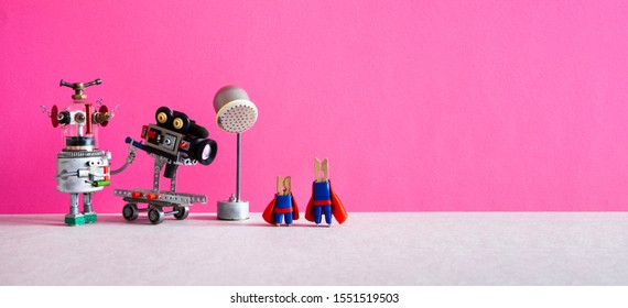 Comical art. Toy robot cameraman makes a movie about super heroes. Funny clothespin actors, robotic operator, camcorder spotlight. Filmmaking behind the scenes concept. Pink background, empty space