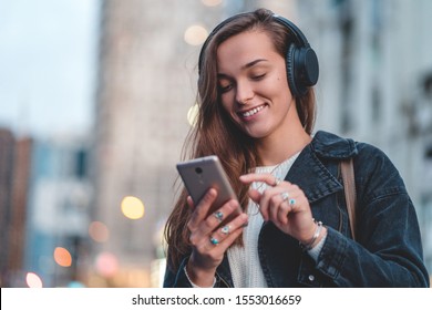 Young happy stylish trendy casual hipster woman changes songs and tracks on smartphone during listening to music on a wireless headphone while walking around the city. Music lover enjoying music