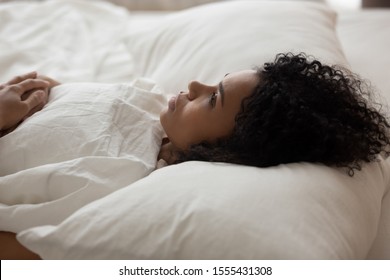 Exhausted African American woman with open eyes lying in bed under white blanket, tired girl with curly hair suffering from insomnia, lack of sleep, early morning awakening concept, close up